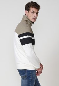 Pánska mikina má vysoký golier a je farebne rozdelená na béžovú, čiernu a bielu. Vyrobená z mäkkej tkaniny, obsahuje rebrované akcenty.