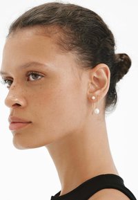 Pendientes de aro de oro con un acento de perla, que presentan un diseño elegante y un acabado pulido. La persona que los lleva tiene un cutis liso y natural.