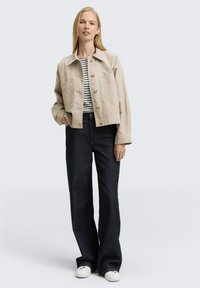 Beige cropped jacket with front pockets, button closure, paired with dark blue flared jeans and white sneakers. Vertical striped shirt underneath.
