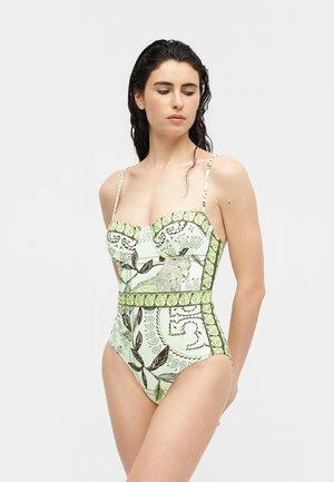 Femeie purtând un costum de baie întreg, verde pal, cu model și bretele subțiri, decorat cu motive botanice, stând în fața unui fundal simplu.