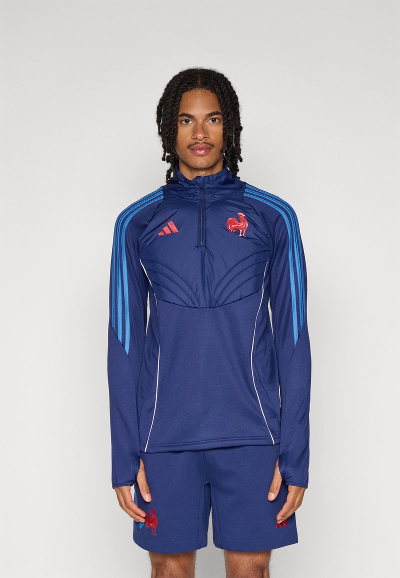 Veste de sport marine avec des rayures bleues et des accents rouges, dotée d'une fermeture éclair à demi, de manches longues et d'un emblème de coq rouge. Associée à un short marine.
