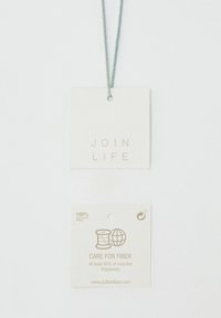 Vierkante labels met groene draad. Het bovenste label leest "JOIN LIFE" in lichte reliëf. Het onderste label vermeldt "CARE FOR FIBER" met een recycling symbool, op een beige achtergrond.