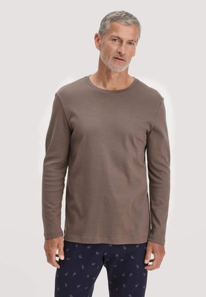 Ein Mann mittleren Alters mit kurzem grauem Haar trägt ein schlichtes braunes Langarmshirt und marineblaue Hosen mit kleinem Paisley-Muster.