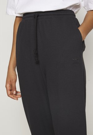 Gros plan sur une personne portant un pantalon de survêtement côtelé noir Adidas avec une taille à cordon de serrage et des poches latérales, associé à une chemise blanche.