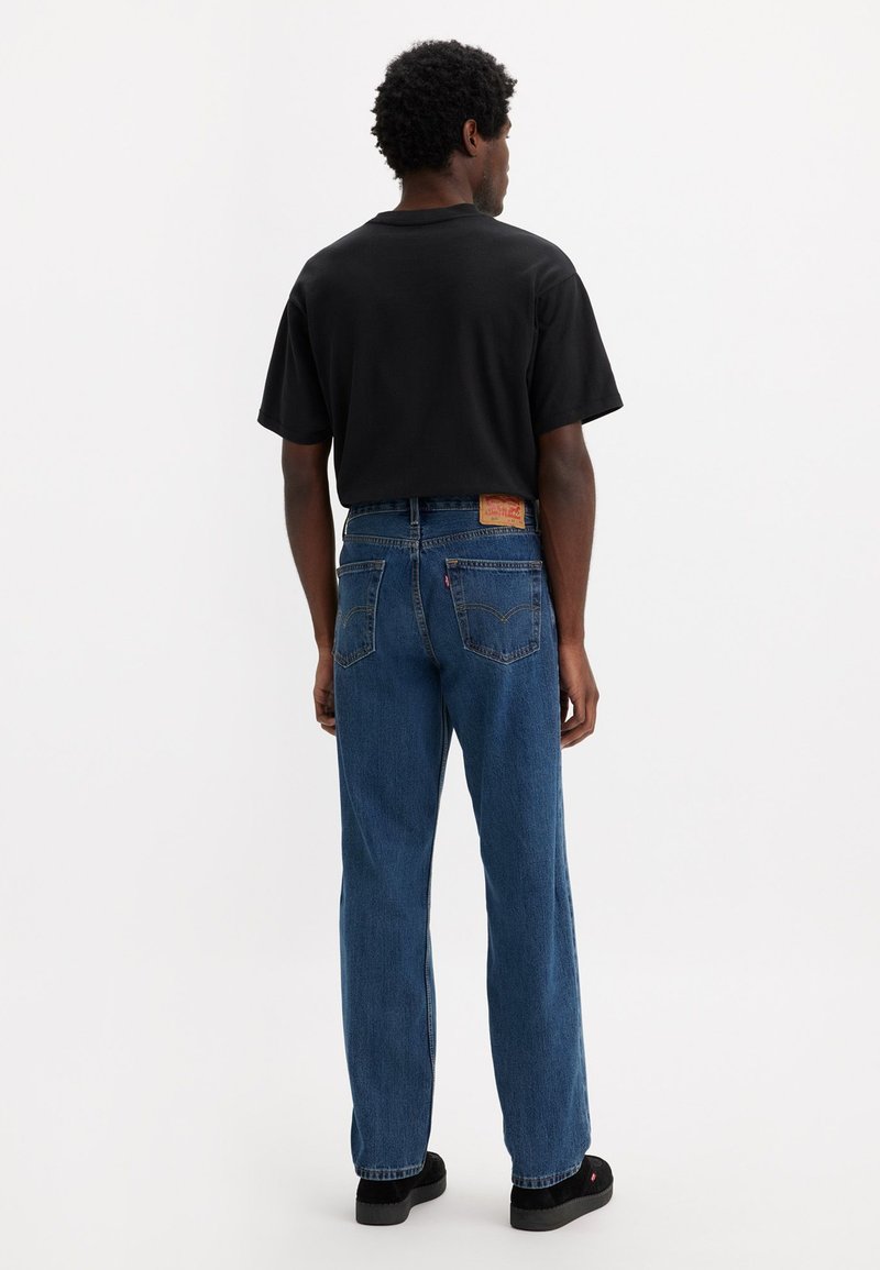 Schwarzes T-Shirt mit kurzen Ärmeln, kombiniert mit blauen Jeans mit geradem Bein, versehen mit einem Lederlogo-Patch am hinteren Bund und dunklem Schuhwerk.