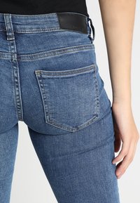 Úzké modré džíny z denimu s zadní kapsou, kontrastním šitím a černou koženou visačkou na pásku.