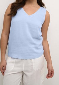 Haut sans manches bleu clair avec un design en V, fabriqué dans un tissu doux, offrant une coupe décontractée et une texture lisse. Associé à un pantalon blanc.