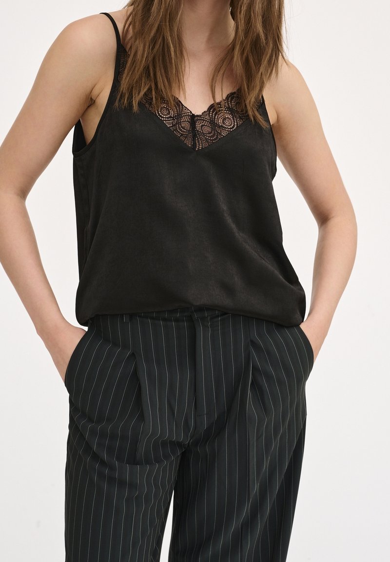 Femme portant un caraco noir bordé de dentelle, rentré dans un pantalon noir à fines rayures, les mains dans les poches, sur fond uni.
