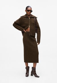 Brun strikket cropped cardigan med glidelås og matchende midiskjørt, med kabelmønster. Akkompagneres med mørke solbriller og hæler.