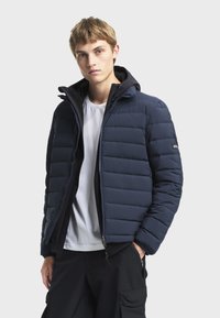 Jeune homme portant une doudoune bleu marine ouverte sur un t-shirt blanc et un pantalon noir, debout devant un fond clair uni.