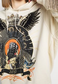 Sudadera con capucha beige con un estampado gráfico que muestra un micrófono retro, alas y un pájaro. Destacada con detalles coloridos y adornos brillantes.