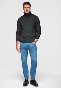 Pull à col roulé gris foncé avec poignets côtelés, associé à un jean bleu clair. Le modèle porte des chaussures en cuir noir et une montre-bracelet.