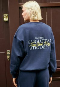 Vaalea nainen, joka on pukeutunut tummansiniiseen huppariin ja housuihin, seisoo selkä kameraan päin ruskean oven edessä. Hupparissa lukee "Manhattan New York City."