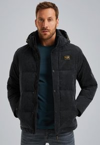 Veste matelassée gris foncé avec une texture douce, capuche, fermeture éclair à l'avant, boutons-pression et un patch logo sur la poitrine. Portée par-dessus une chemise bleue.