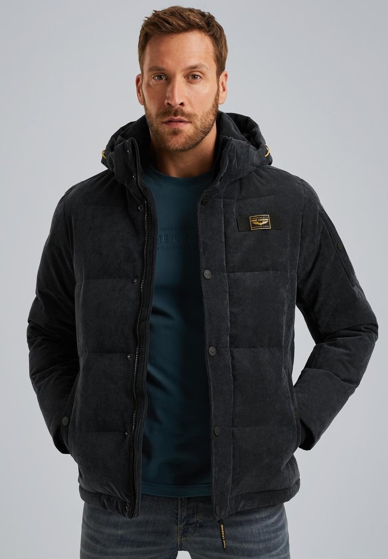 Veste matelassée gris foncé avec une texture douce, capuche, fermeture éclair à l'avant, boutons-pression et un patch logo sur la poitrine. Portée par-dessus une chemise bleue.