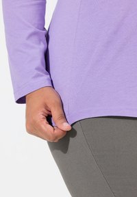 Světle fialový top s dlouhým rukávem a hladkým povrchem; ruka se drží na lemu. Šedé elastické legíny viditelné pod ním.