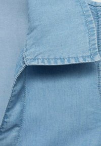 Svijetloplavi denimski materijal s vidljivim ovratnikom, glatka tekstura i uredno šiven rub. Jasne šavove i detalje tkanine su istaknuti.