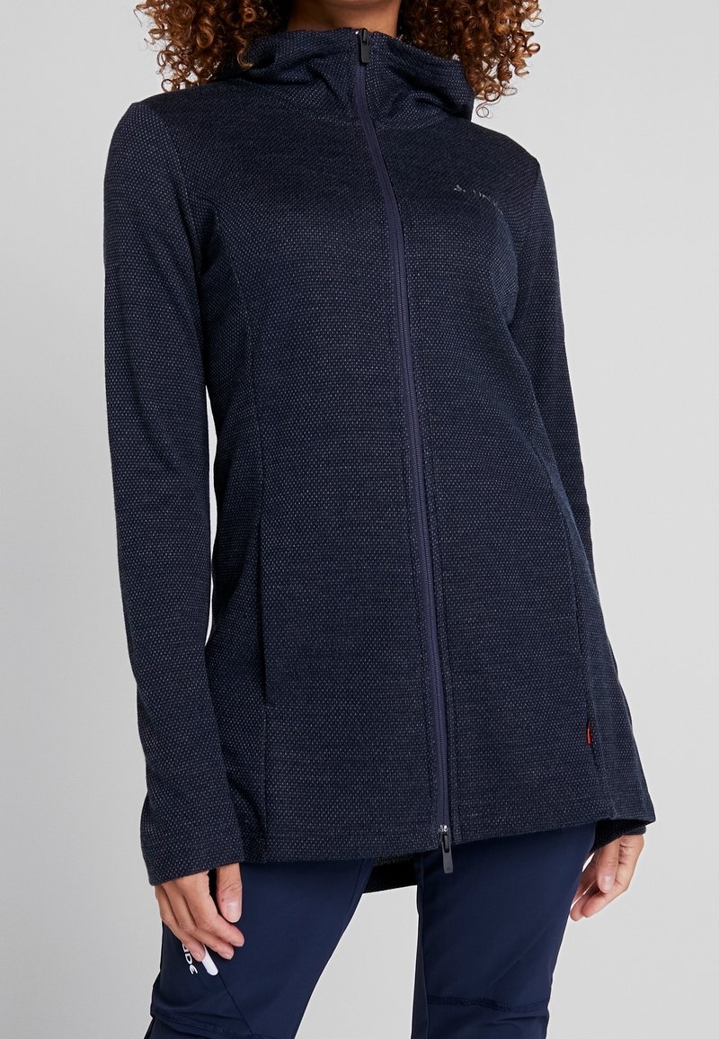 Personne aux cheveux bouclés portant un sweat à capuche zippé texturé bleu marine foncé avec capuche et pantalon bleu marine sur un fond uni.
