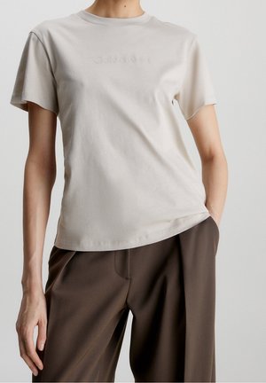 Persoon draagt een beige korte mouw Calvin Klein T-shirt en een losse bruine broek, staand met één hand in de zak.