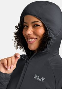 Zwarte hoodie met een ritssluiting, met een gladde textuur en een logo op de borst. Het model heeft krullend haar en glimlacht.