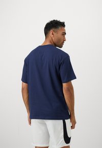 Mand vendt på siden iført en marineblå t-shirt og hvide shorts med et sort swoosh-design på en plain lys baggrund.