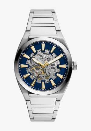 Roestvrijstalen horloge met een blauwe wijzerplaat, gouden indices en een skeleton ontwerp. Voorzien van een zilveren armband en een kroon aan de rechterzijde.