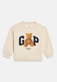 Krēmkrāsas toddler džemperis ar rotaļu lāča attēlu starp lieliem, strukturētiem tumši ziliem burtiem "G" un "P" un zem tā vārdu "Original".