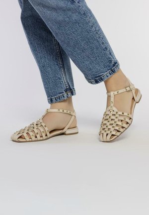 Sandales tissées beiges à bout fermé, avec des accents cloutés et une bride de cheville, associées à un jean cropped bleu. Design à semelle plate.