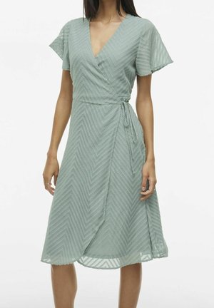 Kobieta w zielonej sukience kopertowej do kolan z krótkimi rękawami i teksturowanym wzorem w jodełkę, zawiązana w talii na bok kokardą.