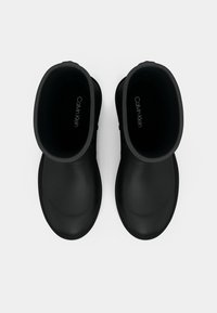 Par črnih Calvin Klein škornjev za dež, pogledanih od zgoraj na beli podlagi, ki prikazuje zaobljene prste in višino do sredine tele.