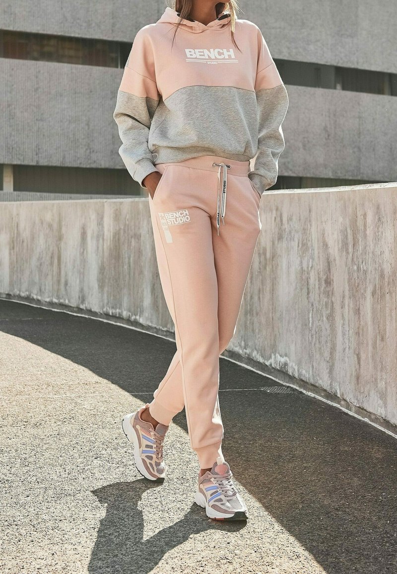 Personne portant un sweat à capuche Bench bicolore rose et gris et un jogging rose, debout à l’extérieur près d’un mur en béton incurvé.