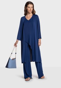 Conjunto fluido de tres piezas en azul marino que incluye una blusa suelta, pantalones de pierna ancha y un cárdigan largo. La modelo sostiene un bolso azul claro.