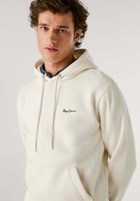 Beige hoodie met een kangoeroezak, een capuchon met trekkoord en een geborduurd logo. Zachte textuur met ribgebreide manchetten en zoom.