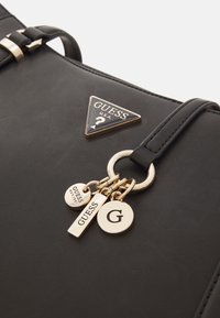 Black synthetic handbag with a smooth texture, featuring gold hardware and three pendants: "G," "GUESS," and "EST. 1981." Logo triangle on the front.