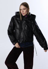 Vrouw in een zwarte pufferjack met capuchon en trekkoorden, donkere broek en een blauwe denim tailleband, poserend met één hand op de heup.