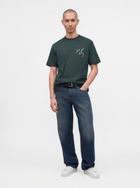 Homme portant un t-shirt vert foncé avec un texte blanc, un jean bleu ample, une ceinture noire et des baskets blanches, debout avec une main dans la poche.