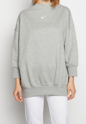 Femme portant un sweat-shirt gris ample Nike avec un petit logo blanc sur la poitrine, assorti à un pantalon blanc, debout devant un fond uni.