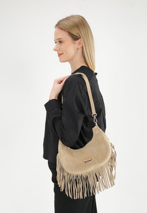 Blonde vrouw in zwarte blouse met een beige suède schoudertas met lange franjes, staand tegen een effen witte achtergrond.