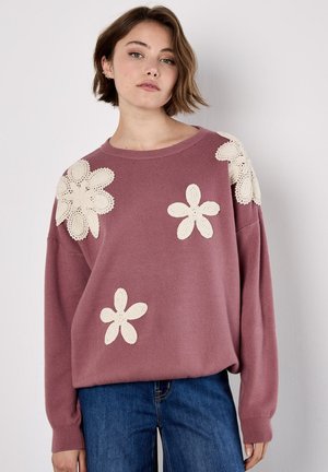 Giovane donna con capelli corti castani che indossa un maglione malva con grandi applicazioni floreali bianche in crochet e jeans di denim blu.