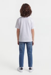 Niño con cabello rizado castaño hasta los hombros, llevando una camiseta blanca de manga corta, jeans azules y zapatillas blancas, de pie y mirando hacia atrás.