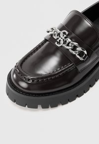 Donkerbruine leren loafer met dikke zwarte zool en zilveren kettinkdetail met ineengestrengelde versierde letters.