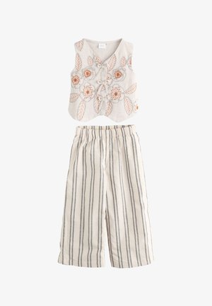 Fată tânără cu păr lung și închis la culoare, purtând un top bej fără mâneci cu broderie florală portocalie și pantaloni în dungi, stând cu o mână în buzunar.