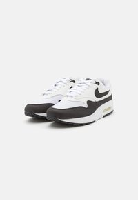 Adidașii Nike Air Max 1 au un superior din textile alb cu accente de suede negru, o unitate de amortizare vizibilă și o talpă exterioară din cauciuc negru.