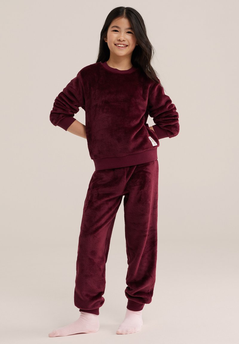 Fille souriante debout, mains sur les hanches, portant un sweat en peluche bordeaux et un pantalon assorti avec des chaussettes rose clair sur fond neutre.