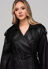 Czarny skórzany płaszcz trench z podwójnym zapięciem, szerokim kołnierzem i paskiem w talii. Posiada pagony na ramionach oraz usztywnioną teksturę.