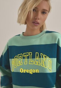 Svetr s pruhy v zelené a námořnické modré barvě s nápisem "PORTLAND" ve žlutém písmu a "Oregon" pod ním, s kulatým výstřihem a měkkou texturou.