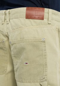 Pantalon en velours côtelé kaki avec un patch en cuir TOMMY JEANS, des accents de boutons en métal et des poches latérales. Présente une surface texturée et une coupe ajustée.