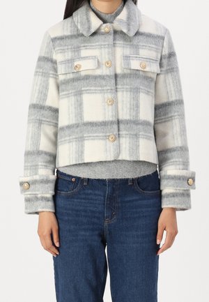 Kvinde iført en grå og hvid ternet uldjakke med knapper over en grå sweater og blå jeans, stående mod en hvid baggrund.