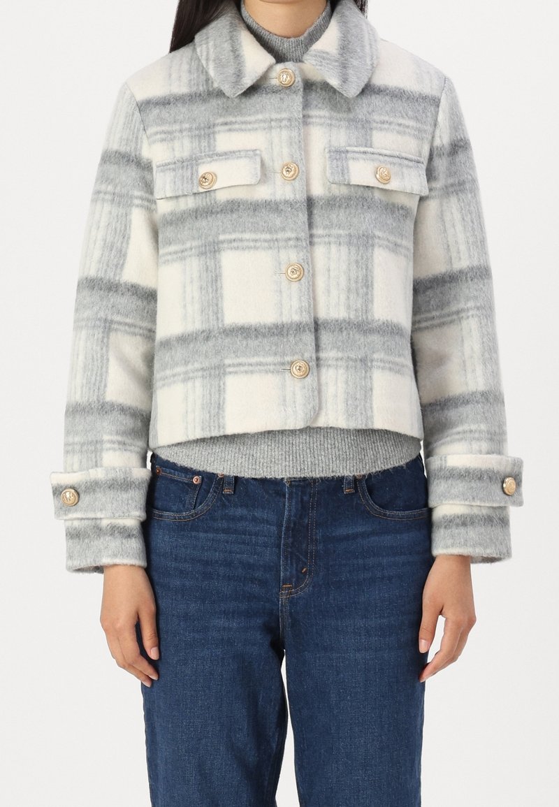 Femme portant une veste en laine à carreaux gris et blanc boutonnée sur un pull gris et un jean bleu, debout sur un fond blanc.