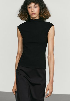 Femme aux cheveux bouclés portant un haut noir sans manches et une jupe noire sur un fond clair uni.